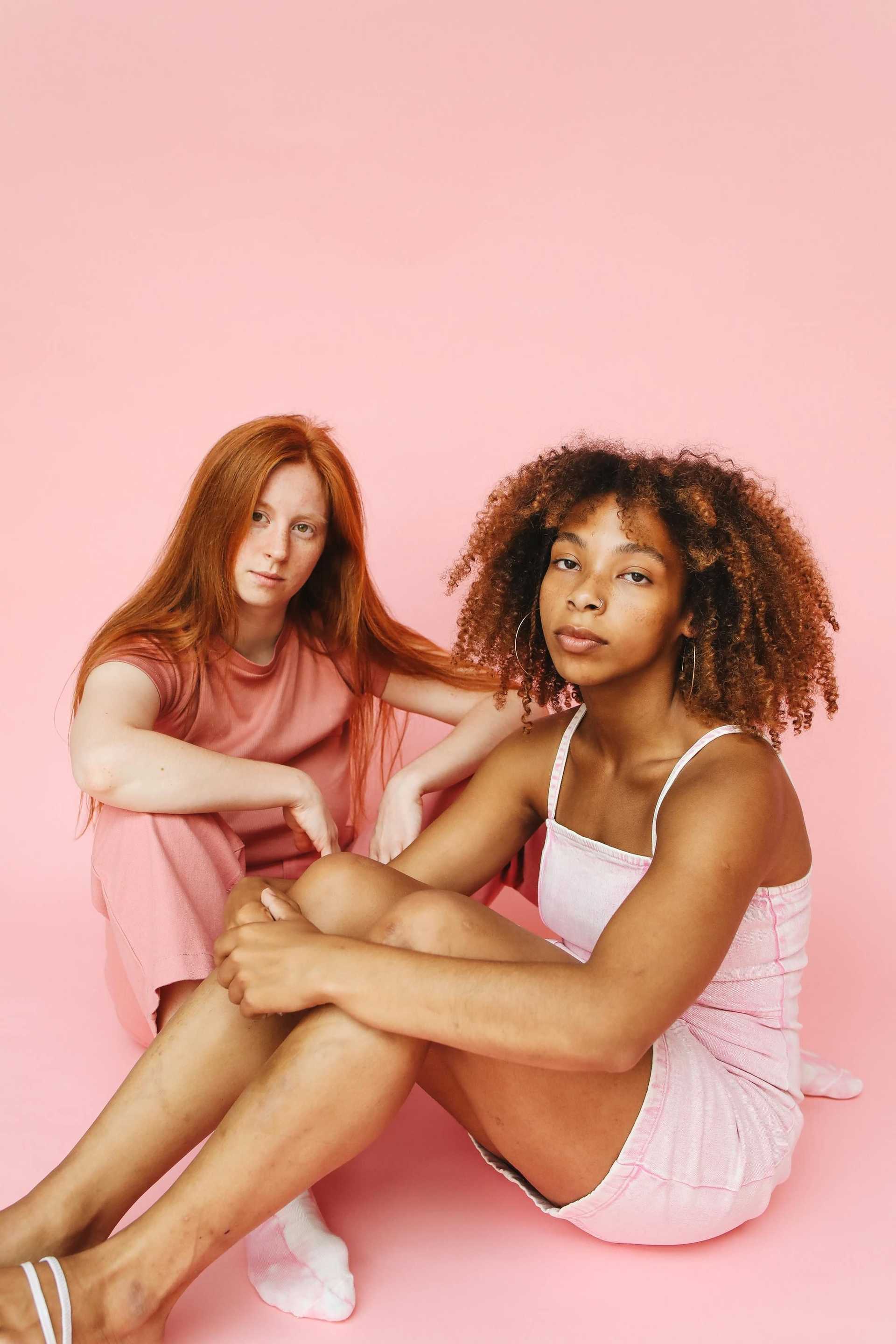 Two women sit against a pink background, wearing casual pink outfits.